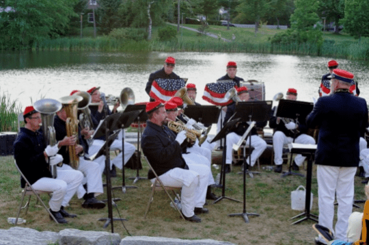 Three rows of brass musicians playing in front of a lake at twilight.